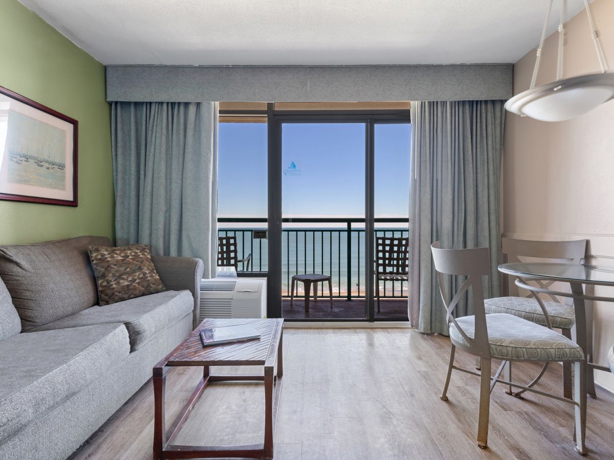 A cozy living room with a gray sofa, a glass-top dining table, chairs, and a large window leading to a balcony with an ocean view.