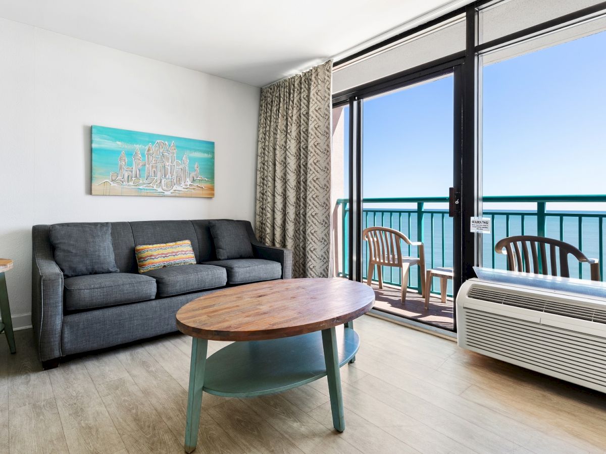 A bright room with a sofa, coffee table, side tables, and a balcony with chairs overlooking the ocean. Art decorates the wall. Air conditioner below.