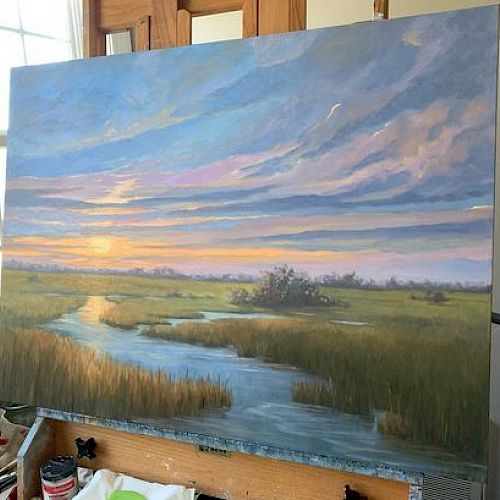 The image shows a painting on an easel, depicting a serene landscape with a winding river, grasslands, and a colorful sunset sky outdoors.