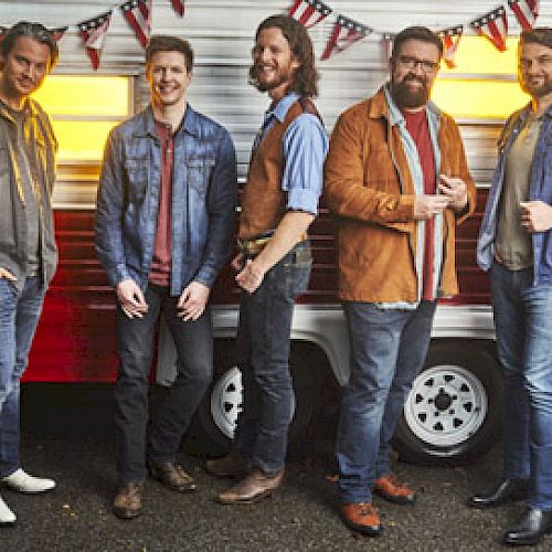 A group of five men are standing in front of a red and white trailer with festive bunting above them. They are casually dressed and smiling.