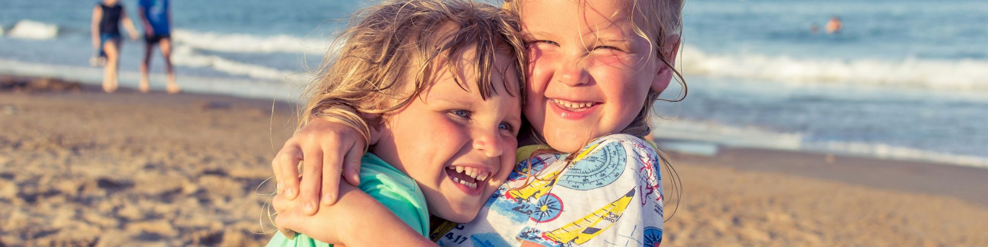 Two children are hugging and smiling on a sandy beach with the ocean in the background. Other people are walking along the shore behind them.