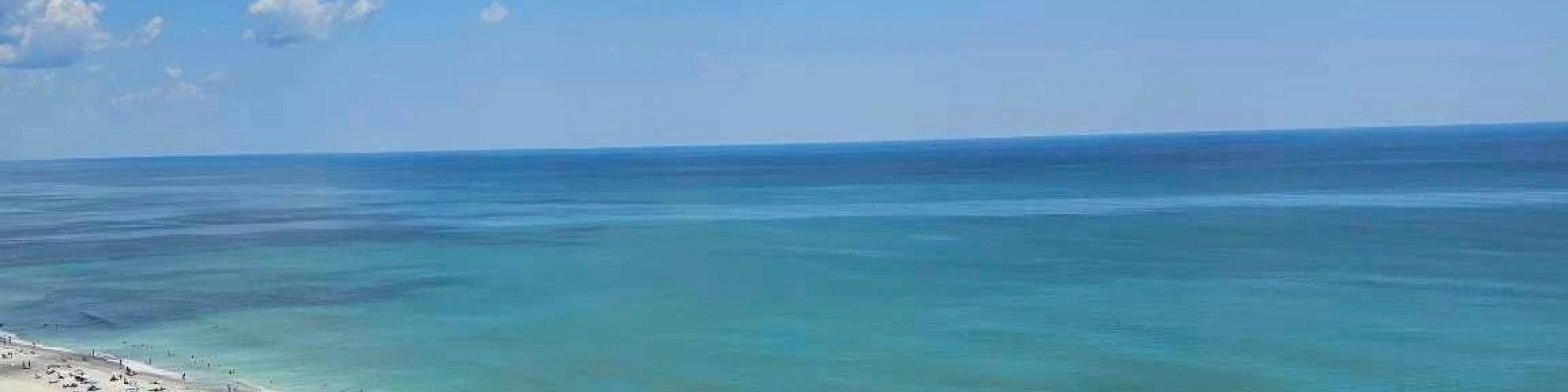 The image shows a beach with clear blue water, people enjoying the sand and sun, colorful umbrellas, and a grassy area with foliage.